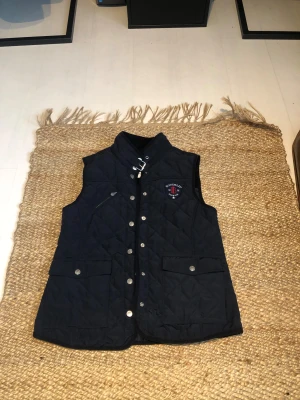 Mörkblå quiltad Bondelid dunväst - Snygg mörkblå quiltad dunväst från Bondelid med silverfärgade knappar, hög krage med spänne och två stora fickor framtill. Västen har ett broderat Bondelid-emblem på bröstet och en normal passform. Perfekt för lager-på-lager under kyliga dagar. Fint skick skriv vid funderingar 