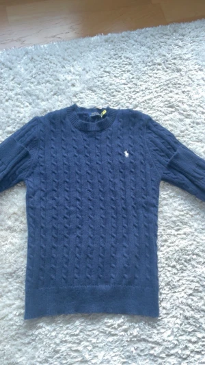 Marinblå stickad tröja Polo Ralph Lauren - Snygg marinblå stickad tröja från Polo Ralph Lauren med klassiskt kabelstickat mönster och vit broderad logga på bröstet. Rund hals och långa ärmar, perfekt för dig som gillar stilrena och tidlösa plagg. Taggarna sitter kvar.