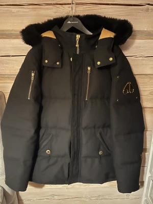 Svart pufferjacka Moose Knuckles - Svart pufferjacka från Moose Knuckles med guldfärgade detaljer och dragkedjor. Jackan har en stor huva med svart fuskpäls, quiltad design och lyxigt guldigt innerfoder med mönster. Snygg logga på ärmen och flera praktiska fickor. Nypris 13 999kr med kvitto.