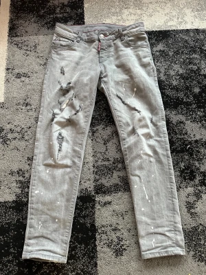 Grå jeans från Dsquared2 med slitningar - Snygga grå jeans från Dsquared2 med coola slitningar och vita färgstänk på både fram- och baksida. Klassisk femficksmodell med röd Dsquared2-tag vid fickan och kontrastsömmar. Jeansen har rak passform och är tillverkade i mjukt bomullstyg.