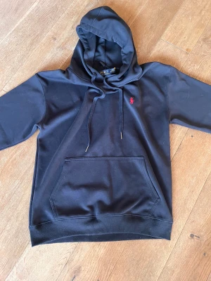 Blå hoodie från Polo Ralph Lauren - Blå hoodie från Polo Ralph Lauren med klassisk röd logobrodering på bröstet. Tröjan har huva med snörning, stor magficka och ribbade muddar. Mjuk bomullskvalitet och normal passform, perfekt för chill dagar.