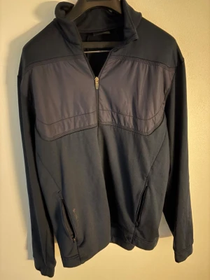 Mörkblå half zip golftröja/golfjacka Galvin Green - Snygg mörkblå långärmad tröja från Galvin Green med half zip och högt krage. Tröjan har två praktiska fickor med dragkedja och diskret logga på ryggen. Materialet är mjukt och känns sportigt, perfekt för golf eller chill dagar. Storlek: L passar M. Nypris: 2999kr