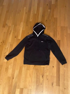 Svart hoodie från BOSS med vita detaljer - Svart hoodie från BOSS med vit kant runt huvan och tunna vita linjer på axlarna. Tröjan har en klassisk känguruficka, snörning i huvan och diskret BOSS-logga på bröstet. Perfekt för dig som gillar stilrena kläder.