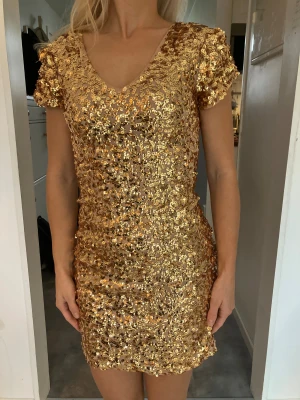 Guldig paljettklänning med v-ringning - Superglittrig kort klänning i guld med täckande paljetter över hela plagget. V-ringad både fram och bak, med korta puffärmar och figurnära passform. Perfekt för dig som vill sticka ut och glänsa på nästa event! Använd en gång. 