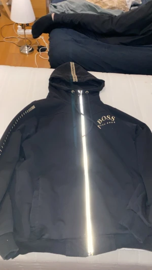 Svart vindjacka från Hugo Boss - Svart kofta  från Hugo Boss med guldiga detaljer och logga på bröstet. Kofta har huva med snörning, hel dragkedja framtill och diskreta mönsterdetaljer på ärmen. Tillverkad i polyester för en sportig och clean look.