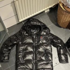 Svart pufferjacka från Polo Ralph Lauren - Svart pufferjacka från Polo Ralph Lauren med glansig finish, huva och flera dragkedjefickor. Jackan har en röd Polo-logga på bröstet och är fodrad för extra värme. Perfekt för kalla dagar och har en modern, loose passform.