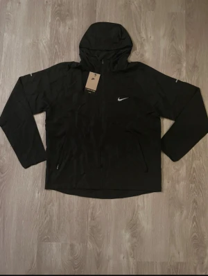 Svart Nike vindjacka med huva men den e liten i storlek så S passar - Svart vindjacka från Nike med huva och dragkedja framtill. Jackan har två sidofickor med dragkedja och en vit Nike-logga på bröstet. Tillverkad i lätt polyester, perfekt för löpning och utomhusaktiviteter. Snygg och sportig design med lång ärm.