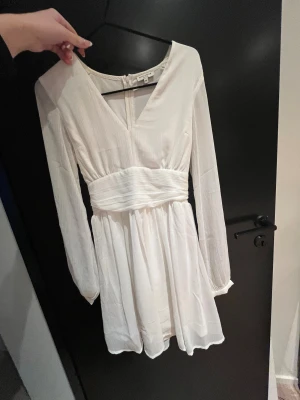 Vit omlottklänning med knyt i midjan - Superfin vit kortklänning från bubbelroom med v-ringning, långa ärmar och knytband i midjan. Klänningen har en lätt, flowy känsla och dragkedja i ryggen. Perfekt för dig som gillar en stilren och fräsch look.