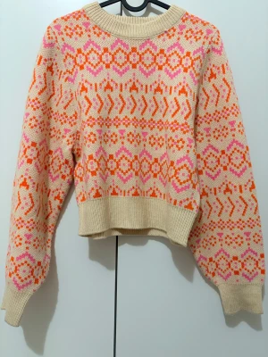 Beige stickad tröja med mönster från H&M - Superfin stickad tröja från H&M i beige med färgglatt mönster i orange och rosa. Tröjan har rund hals, ribbade muddar och långärmade ärmar. Perfekt för dig som gillar retrovibbar och vill ha något som sticker ut i garderoben.