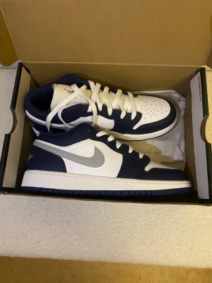 Nike Air Jordan 1 Low marinblå/vit - Nike Air Jordan 1 Low sneakers i marinblått och vitt med grå Swoosh. Klassisk rund tå, snörning och platt sula. Skorna har perforerad tåbox och Air Jordan-logga broderad på hälen. Tillverkade i skinn och syntetmaterial för en clean och sportig look.