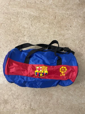 FC Barcelona sportväska blå/röd - Sportväska med FC Barcelona-logga och LFP-märke, i blått och rött med svarta handtag. Ena sidan har en cool fotbollsinspirerad form och tryck. Perfekt för träning eller matchdagar, tillverkad i slitstark polyester.