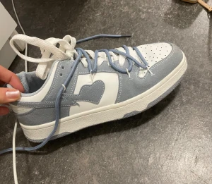 Vita och ljusblå sneakers med hjärtdetalj - Snygga sneakers i vitt och ljusblått med en unik hjärtformad detalj på sidan. Skon har både vita och ljusblå snören.