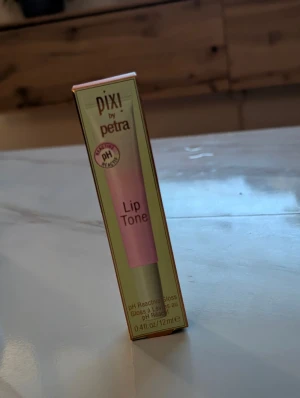 Pixi Lip Tone Prettiness läppglans - Säljer ett Pixi Lip Tone Prettiness läppglans med pH-reaktiv formula som ger en personlig glansig ton. Innehåller nyponfröolja och mangosmör för extra fukt. Tuben är rosa och grön, 12 ml. Perfekt för dig som vill ha en naturlig tint med gloss. Obruten förpackning 