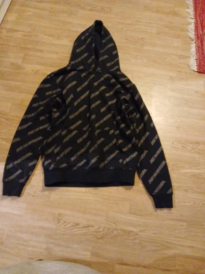 Svart hoodie med text från FSBN - Jag har en till med storlek L.                Svart hoodie från FSBN i storlek M med huva och lång ärm. Tröjan har ett mönster med gul text 'JUST DO NOTHING.' tryckt över hela plagget. Regular fit och tillverkad i bomullsmix, perfekt för chill dagar. Snygg och enkel att matcha med jeans eller joggers.