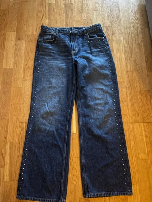 Blå jeans med nitar och wide fit - Snygga blå jeans med breda ben och coola nitar längs sidorna. Klassisk femficksmodell med normal midja och raka sömmar. Jeansen har en mörk tvätt och är tillverkade i robust denim. Perfekt för dig som gillar att sticka ut med detaljer.