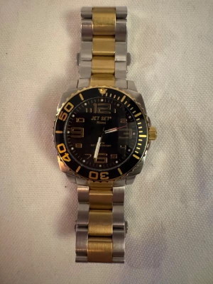 Jet Set Miami klocka i guld och silver - Jet Set Miami herrklocka/unisex med robust länkarmband i guld och silverfärgad metall. Svart urtavla med stora siffror och gulddetaljer, vridbar bezel och tydliga markeringar. Klockan har en sportig och stilren look med metallspänne och Jet Set-logga på baksidan. Klarar av regn och stänk. Inget fungerande batteri i så vet ej om klockan funkar med nytt. Extra delar kommer med 
