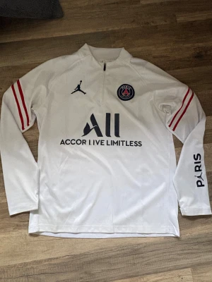 PSG vit långärmad fotbollströja S - Vit långärmad PSG fotbollströja i samarbete med Jordan. Tröjan har svarta tryck, rött randigt mönster på ärmarna, halv dragkedja och Paris Saint-Germain-logga på bröstet. Snyggt tryck på ryggen och ärmen.