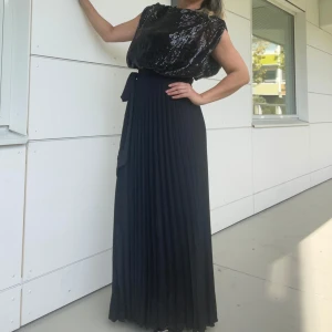 Svart glittrig långklänning - Snygg svart långklänning med glittrig överdel och ett elegant knytband i midjan. Perfekt för festliga tillfällen! Den har en ärmlös design och ett plisserat kjolparti som ger en fin rörelse. Klänningen är både stilren och iögonfallande, perfekt för en kväll ute.Helt ny.Finns i tre storlek S,M och L