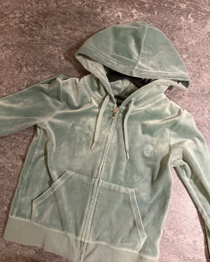 Mintgrön velour hoodie Juicy Couture - Säljer en mintgrön hoodie från Juicy Couture i mjuk velour med dragkedja och snörning vid huvan. Tröjan har långa ärmar, fickor framtill och ett broderat JC-logo på bröstet. Perfekt för chill dagar och har en riktigt skön vibe.