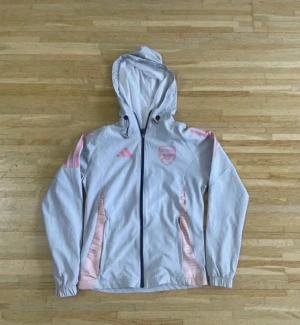 Vit vindjacka Arsenal x Adidas - Snygg vit vindjacka från Adidas i samarbete med Arsenal. Jackan har huva, hel dragkedja och rosa detaljer på axlar och sidor. Perfekt för dig som gillar sportig stil och vill visa ditt lagstöd. Lätt material och klassisk passform.
