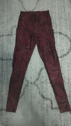 Blommiga vinröda leggings från Gymology XS - Supersnygga vinröda leggings från Gymology i storlek XS, med blommigt mönster och hög midja. De har praktiska sidofickor och är gjorda i mjukt, stretchigt syntetmaterial som sitter tight och bekvämt. Perfekta för träning eller chill.