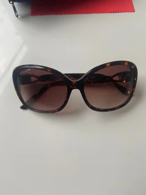 Bruna Dior solglasögon med gulddetalj - Snygga solglasögon från Dior med brunmelerad båge i kattögonform och stora glas. Sidorna har guldfärgade detaljer och logotypen på skalmen. Perfekt accessoar för att lyfta din stil och ge en lyxig känsla.