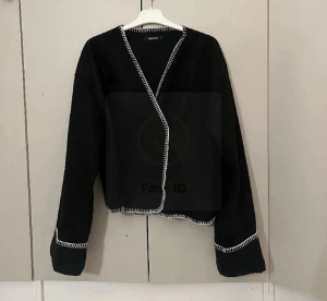 Svart omlottkofta från Ginatricot - Svart omlottkofta från Ginatricot med vita dekorativa sömmar längs kanterna och ärmsluten. Jackans ena knapp är av och den andra finns kvar men funkar ej. Men man kan ha den som en kofta osv, strl men sitter som en S eller m. 