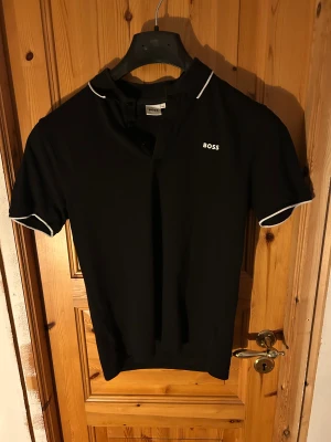Svart pikétröja från BOSS med vita detaljer - Svart pikétröja från BOSS i storlek 164 men passar runt 170 också med vit logga på bröstet och vita kantdetaljer vid krage och ärmslut. Klassisk design med korta ärmar och knappar vid halsen. Perfekt för dig som gillar stilrena och snygga plagg. Priset kan diskuteras