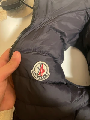 Svart Moncler pufferjacka S - Säljer en svart pufferjacka från Moncler i storlek S. Jackan har klassisk quiltad design, dragkedja framtill och Moncler-logga på bröstet. Materialet är glansigt och lätt, perfekt för kyliga dagar. Insidan har ett unikt serietidningsmönster med tvättråd.
