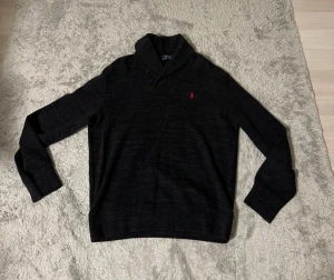 Svart stickad cardigan från Polo Ralph Lauren - Snygg svart polotröja cardigan från Polo Ralph Lauren med klassisk röd logobrodyr på bröstet. Tröjan har lång ärm, ribbade muddar och en stilren krage med knappar. Perfekt för dig som gillar en clean och tidlös look.