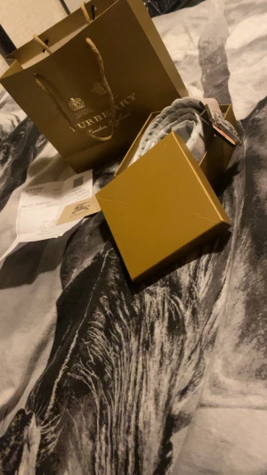 Rutig halsduk från Burberry - Säljer ett klassiskt bälte från Burberry med ikoniskt mönster i beige, vit, röd och grå. Bältet har en snygg metall-detalj med Burberrys logotyp. Perfekt accessoar för att lyfta din stil och levereras i originalförpackning.