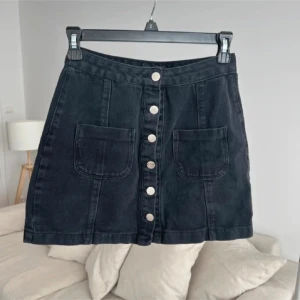 Svart jeanskjol från I Saw It First - Svart kort jeanskjol med raka linjer, stora framfickor och silvriga knappar längs hela kjolen. Den är lite urtvättad i färgen