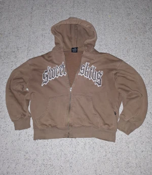 Brun hoodie Sweet Sktbs - Brun hoodie från Sweet Sktbs med stor vit och grå text på bröstet. Tröjan har dragkedja, huva och känguruficka. Tillverkad i mjuk bomullsmix, perfekt för chill dagar. Pris kan diskuteras