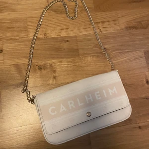 Vit axelväska från Carlheim med kedja - ⚠️SKRIV TILL MIG INNAN DU KÖPER, då den ligger ute på andra ställen också⚠️ Snygg vit axelväska från Carlheim med stilren design och guldfärgad kedjerem. Remen går att ta av om man skulle vilja det.  Väskan har ett lock med tryckknapp och diskret logga framtill. Perfekt storlek för det viktigaste och passar till många outfits. Bredd: 22cm höjd: 13cm. Inte superkvalitet därav priset. Det är några fläckar från ”locket” se bild 3.