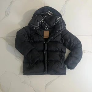 Svart pufferjacka från Burberry - Svart pufferjacka från Burberry med huva och dragkedja framtill. Jackan har en stor logopatch på ärmen och klassisk Burberry-detalj på dragkedjan. Fodret är quiltat och jackan är perfekt för kalla dagar. Snygg och stilren design med en loose passform. Jackan värmer ganska snabbt och är för dig som söker en jacka med både stil och värme under vintertiderna. Kan också användas vid sommartider då man kan dra bort ärmarna så att de blir till en väst istället. Pris kan diskuteras 