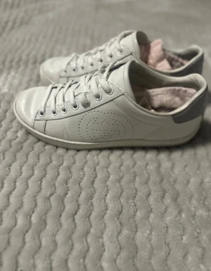 Vita sneakers från Gucci i skinn - Snygga vita sneakers från Gucci i skinn med klassisk låg profil och rund tå. Skorna har snörning och diskret perforerat GG-logo på sidan samt grå detalj vid hälen. Perfekta för dig som gillar stilrena och lyxiga sneakers.