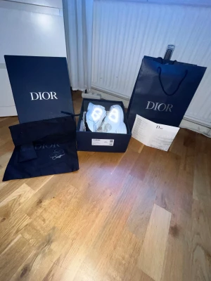 Vita Dior sneakers med reflekterande detaljer - Säljer ett par exklusiva vita Dior sneakers med reflekterande CD-detaljer på sidorna. Skorna har snörning, rund tå och platt sula. Materialmix av skinn och syntet med meshpaneler för extra komfort. Kommer med originalkartong, dustbag och äkthetsintyg.