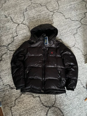 Svart pufferjacka från Polo Ralph Lauren - Svart pufferjacka från Polo Ralph Lauren med huva och dragkedja framtill. Jackan har ett rött Polo-logo broderat på bröstet, två sidofickor med dragkedja och quiltad design. Materialet är glansigt och syntetiskt, perfekt för kalla dagar.