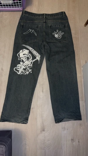 Grå jeans med tryck - Unika grå jeans med bred passform och coola vita tryck på bakfickorna och benet, bland annat en reaper-illustration och grafiska detaljer. Klassisk femficksmodell med knapp och dragkedja. Materialet är robust denim som ger en snygg streetkänsla.