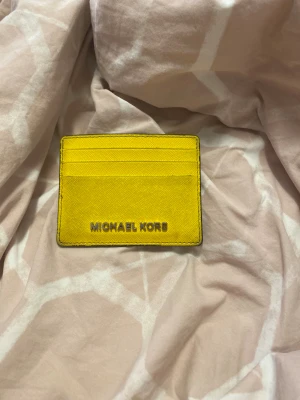 Gul korthållare från Michael Kors - Snygg gul korthållare från Michael Kors i skinn med flera kortfack och ett fönster för ID på baksidan. Perfekt att ha i fickan eller väskan när du vill ha med dig det viktigaste. Stilren design med silverfärgad logga framtill.