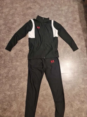 Svart träningsset från Under Armour - Säljer ett svart träningsset från Under Armour i storlek S. Setet består av en zip-jacka med vita paneler på axlarna och matchande byxor. Båda delarna har rött Under Armour-logga. Materialet är mjukt och stretchigt, perfekt för träning eller sport.