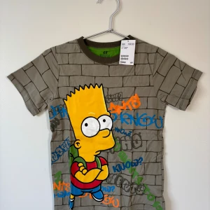  Bart Simpson t-shirt - 134/140 - En t-shirt från med grått tegelstensmönster och stor färgglad Bart Simpson-tryck på framsidan. T-shirten har korta ärmar och graffitiinspirerade detaljer i orange, blått och grönt. 