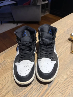 Nike Air Jordan 1 svart/blå sneakers - Säljer ett par Nike Air Jordan 1 sneakers med svart, vit och blå färg. Skorna har högt skaft, klassisk snörning och rund tå. Swoosh-loggan i blått på sidan och Air Jordan-märket på ankeln. Materialet är skinn och syntet, med slitstark platt sula.