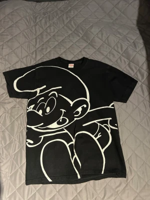 Supreme Smurfarna svart t-shirt - Supreme t-shirt i svart bomull med stor vit Smurfarna-grafik på framsidan och SUPREME-tryck över ryggen. Klassisk passform och korta ärmar, perfekt för dig som gillar streetwear och unika prints.
