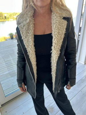 Svart skinnjacka med teddykrage Urban Code - Snygg svart skinnjacka från Urban Code med krämvit teddyfoder och coola quiltade detaljer på ärmarna. Jackan har dragkedja framtill och en längre, figurnära passform. Perfekt för dig som vill ha en edgy look med extra värme.