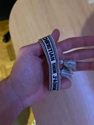 Christian Dior J'Adior armband - Snyggt vävt armband från Christian Dior med texten 'J'Adior' i svart på beige botten. Armbandet har justerbar knytning med tofsar i ändarna och en cool, casual vibe. Perfekt accessoar för att lyfta din outfit med en touch av lyx.