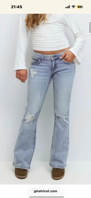 Ljusblå bootcut jeans med slitningar - Säljer ett par ljusblå bootcut jeans från Gina Tricot med låg midja och snygga slitningar på låren och knäna. Jeansen har klassiska fem fickor, markerade sömmar och dekorativa ficklock bak. Perfekt för dig som gillar retrovibbar och coola detaljer.