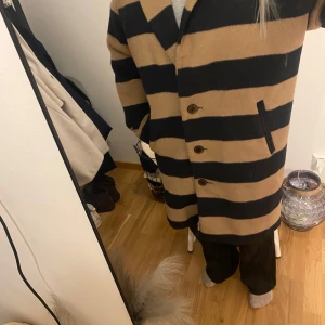 Randig kappa från Lindex  - Säljer en snygg randig kappa från Holly and whyte beige och svart. Jackan har breda horisontella ränder, klassisk krage och tre knappar framtill. Två fickor med sned öppning . Ena innertyget på fickan behöver sys 