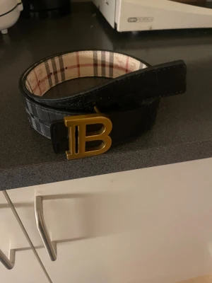 Burberry bälte med guldspänne - Snyggt Burberry bälte med klassiskt rutigt mönster i beige, svart och rött. Bältet har ett stort guldigt B-spänne och är gjort i skinn. Perfekt accessoar för att lyfta din outfit med en lyxig touch.