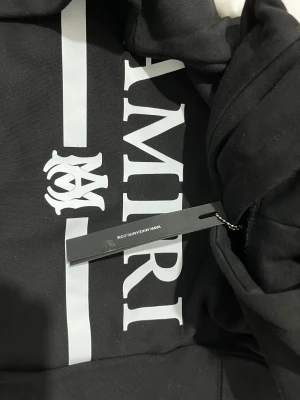 Svart AMIRI hoodie med vit logga - Svart hoodie från AMIRI med stor vit logga och emblem på bröstet. Tröjan har huva, magficka och ribbade muddar. Snygg och stilren design, perfekt för dig som gillar streetwear och exklusiva märken.